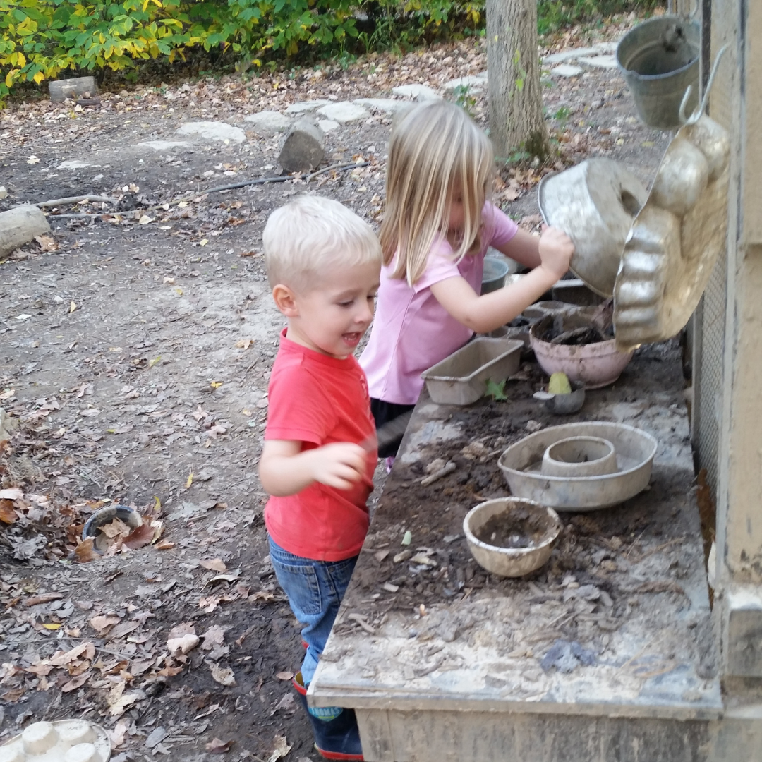 A blonde boy in a red shirt and a blonde girl in a pink shirt play at the mud pie station in the Mud Zone at the Nature PlayScape